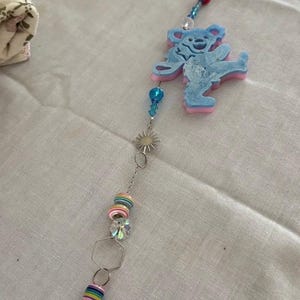 May include: A decorative suncatcher featuring a blue dancing bear charm with pink accents, strung with a silver chain and colorful beads. The suncatcher also includes a blue and teal agate stone pendant.