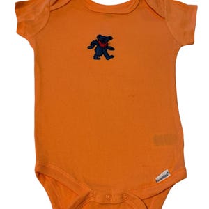 May include: Bright orange baby bodysuit featuring a blue and red bear emblem. The short-sleeved bodysuit has poppers at the bottom. The brand name "onesies" is visible on the lower right side.