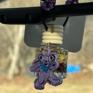 Puede incluir: Encanto de oso morado y azul colgando de una botella de vidrio transparente llena de flores secas. La botella está unida a un difusor de cuentas de madera blancas.