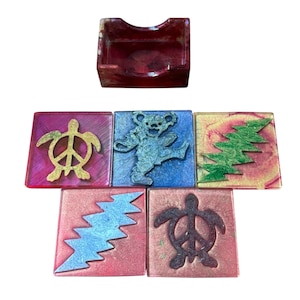 May include: Set of six colourful coasters with various designs, including a peace sign with a turtle, a dancing bear, and a lightning bolt. The coasters are made of resin and have a glossy finish. A matching coaster holder is also included.