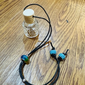 Puede incluir: Una cuerda negra con dos cuentas azules y verdes y una pequeña botella de vidrio con una tapa de madera y un dije de hoja. La botella cuelga de la cuerda.