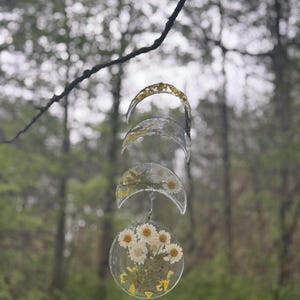 May include: A hanging mobile with five clear resin crescent moons and a round resin circle with pressed white flowers and green leaves. The mobile is hanging from a branch in a forest.