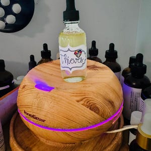 May include: A clear glass bottle with a black dropper, labeled "Thieves," sits atop a wooden essential oil diffuser. The diffuser has a purple light and the brand name "InnoGear" is visible. Other amber bottles are in the background.