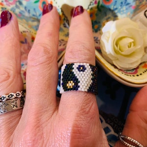 May include: A white and black beaded ring with gold accents. The ring is on a person's finger.