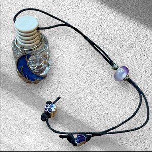 Puede incluir: Una cuerda negra con una botella de vidrio transparente con un diseño de luna creciente azul, una cuenta morada, un dije de mariposa y un dije de corazón.