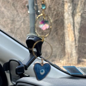 Puede incluir: Un amuleto de coche azul y dorado con un diseño de ojo turco en forma de corazón que cuelga del espejo retrovisor.