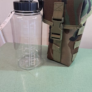 May include: A clear plastic water bottle with a black lid and markings for measuring liquid in ounces. The bottle is next to a camouflage-patterned water bottle carrier.