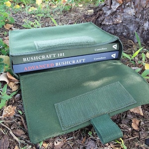May include: A green canvas book bag with a flap and a strap. The bag is open and two books are inside. The books are titled "Bushcraft 101" and "Advanced Bushcraft".