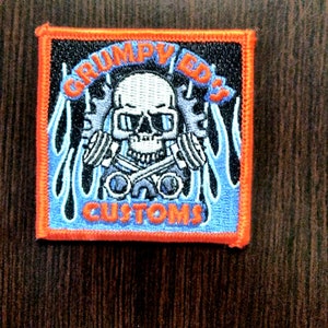 Puede incluir: Un parche bordado negro y azul con una calavera con gafas de sol y sosteniendo llaves inglesas. La calavera está rodeada de llamas y el texto "Grumpy Ed's Customs" está escrito debajo.