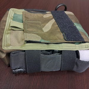 May include: Camouflage-patterned tactical medical pouch with black straps and a black loop. The pouch has a flap secured with a hook-and-loop fastener. A white bandage is visible in a side pocket.