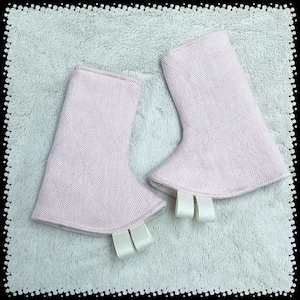 May include: A pair of light pink fabric boot covers with a diamond pattern. Each cover has a white ribbon tag attached at the bottom. The covers are displayed on a light gray, textured surface.