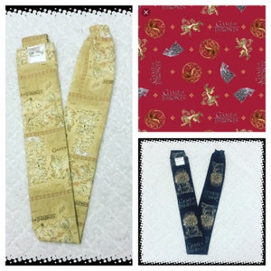 May include: Two fabric headbands with a Game of Thrones theme. One headband is a beige color with a map of Westeros and the text "Game of Thrones". The other headband is a dark blue color with a gold dragon and the text "Game of Thrones".