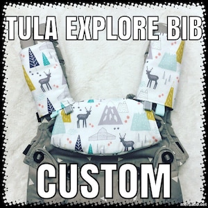 Peut inclure: Un bavoir personnalisé Tula Explore pour porte-bébé avec un fond blanc et un motif de cerfs, de montagnes et d'arbres en gris, sarcelle et jaune. Le texte "TULA EXPLORE BIB CUSTOM" est affiché en lettres blanches.