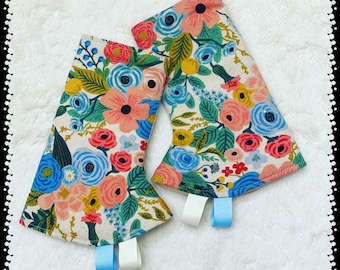 May include: A pair of floral print oven mitts with blue and white ribbon ties. The mitts are made of a light-colored fabric with a floral pattern in shades of pink, blue, green, and yellow.