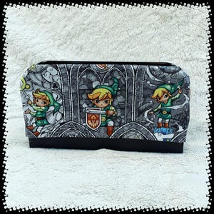 May include: A rectangular, black-bottomed case with a repeating pattern of a cartoon character in a green tunic and hat, holding a sword and shield. The background is a stained-glass window design in shades of gray and white.
