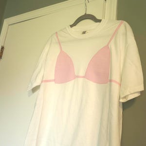 Puede incluir: Una camiseta blanca de manga corta y cuello redondo con un estampado gráfico de un top de bikini con patrón de cuadros vichy en rosa y blanco en la parte delantera. El diseño incluye tirantes finos rosas que se extienden hacia los lados.