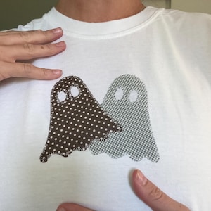 May include: A white t-shirt featuring two ghost-shaped fabric appliqués stitched onto the front. The left ghost is brown with white polka dots, and the right ghost is a grey and white checkered pattern. Both ghosts have two oval eye cutouts.