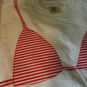 May include: A white crew neck t-shirt featuring a red and white horizontal striped bikini top graphic appliqued onto the front. The graphic is stitched with a visible zigzag pattern along the edges. The shirt has a Comfort Colors brand label.