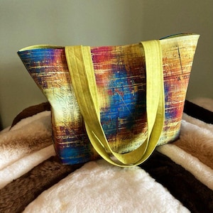May include: A colorful tote bag with a map-like design in shades of blue, red, and yellow. The bag has bright yellow handles and a matching interior lining. The bag is sitting on a soft, textured surface.
