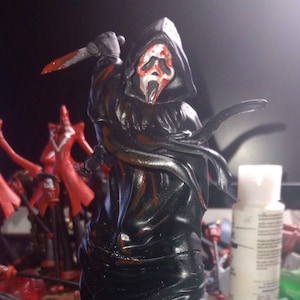 Könnte beinhalten: Eine schwarz und rot bemalte Ghostface-Figur aus der Scream-Filmreihe. Die Figur hält ein Messer und steht auf einem dunkelvioletten und schwarzen Sockel.