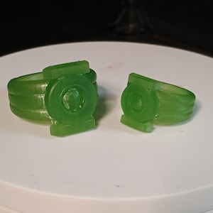 Może przedstawiać: Dwa przezroczyste zielone pierścienie, każdy z okrągłym emblematem i prostokątną podstawą. Pierścienie mają warstwowy wzór opaski. Jeden pierścień jest kompletny, drugi jest częściowy.