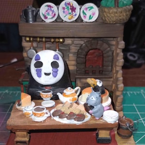 May include: A No Face character from the movie Spirited Away sits at a table with a small grey cat-like creature. The table is set with teacups, saucers, a teapot, a plate of cookies, and a plate of cheese. The scene is set in front of a brick fireplace with a basket of green leaves on top.