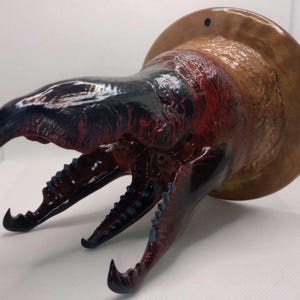 May include: A dark red and black ceramic sculpture of a creature's head with a large, open mouth and sharp teeth. The sculpture is mounted on a round, wooden base.