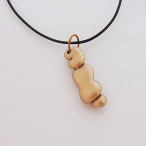 Può includere: Una collana con un ciondolo dorato di forma organica astratta unica. Il ciondolo è realizzato in un materiale liscio e arrotondato e pende da un cordoncino nero.