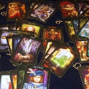 May include: A collection of tarot cards with various illustrations, including figures, symbols, and scenes. The cards are spread out on a dark blue surface, with some cards featuring the words "Swords", "Wands", and "Page of Swords".