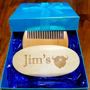 May include: A light wood beard brush and comb set inside a blue gift box. The oval brush is engraved with "Jim's" and a mustache design. The box is tied with a blue ribbon.