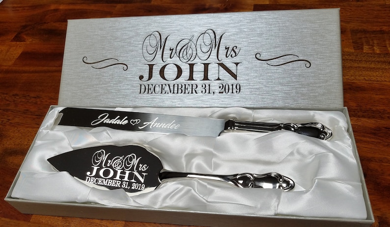 May include: A silver wedding cake knife and server set in a white satin lined box. The knife and server are engraved with "Mr. & Mrs. John December 31, 2019". The knife is also engraved with "Jadale & Anndee".