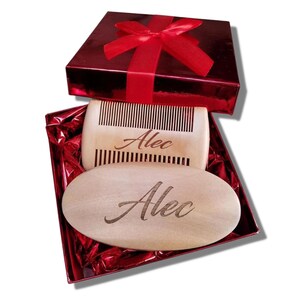 May include: A wooden comb and oval-shaped beard brush, both engraved with the name "Alec", are presented inside a red gift box with a matching ribbon. The comb has vertical teeth, and the brush is smooth. The gift box is a shiny red.