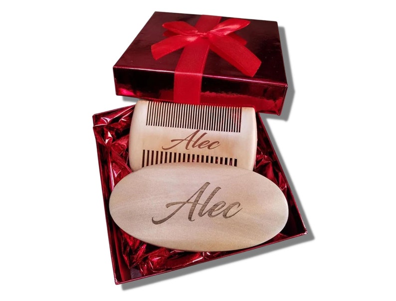 May include: A wooden comb and oval-shaped beard brush, both engraved with the name "Alec", are presented inside a red gift box with a matching ribbon. The comb has vertical teeth, and the brush is smooth. The gift box is a shiny red.
