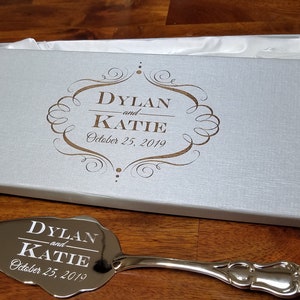 May include: A silver cake server with a decorative handle and a mirrored surface engraved with "Dylan and Katie October 25, 2019" in a white box with a gold inscription.