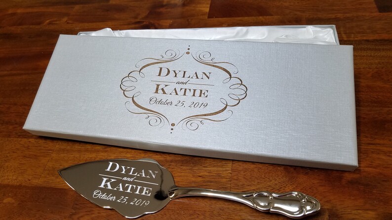 May include: A silver cake server with a decorative handle and a mirrored surface engraved with "Dylan and Katie October 25, 2019" in a white box with a gold inscription.