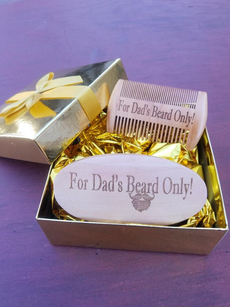 May include: A gold gift box with a yellow ribbon contains a wooden beard comb and a wooden beard brush. The comb and brush are engraved with the text "For Dad's Beard Only!" and a beard graphic. The box is lined with gold tissue paper.