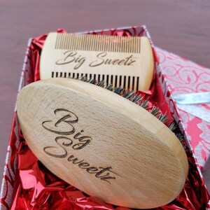 May include: A wooden beard brush and comb set in a gift box. The brush is oval-shaped with bristles, and the comb has fine teeth. Both items are engraved with the words "Big Sweetz". The box is red with a silver pattern.