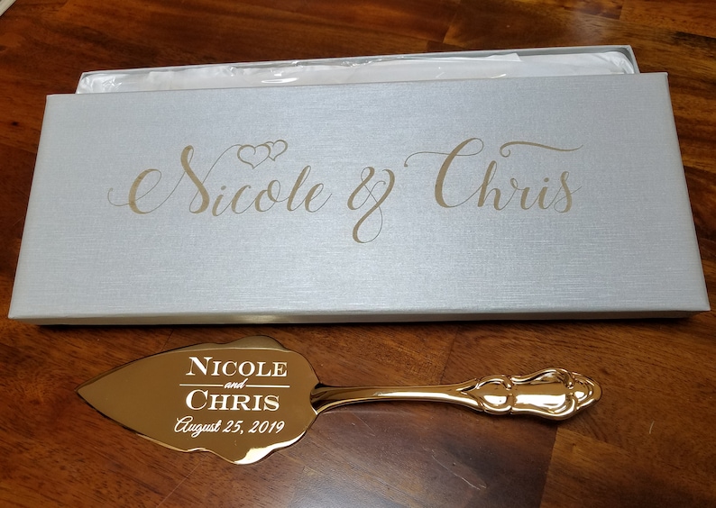 May include: Gold cake server with a leaf-shaped blade engraved with "Nicole and Chris August 25, 2019". The server comes in a white box with the names "Nicole & Chris" engraved in gold.
