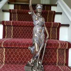 May include: A bronze statue of the figure of Justice, blindfolded, holding a sword and scales, standing on a book with a snake. The statue is on a black base with the text "LCTR, PLLC Est 2019".