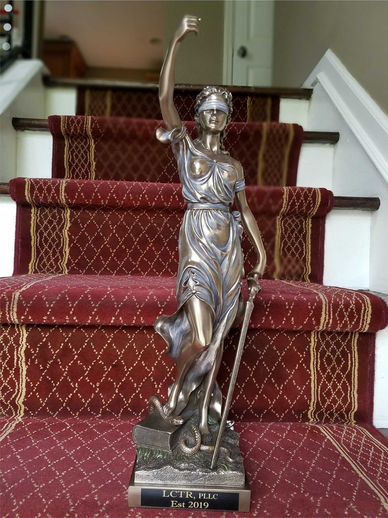 May include: A bronze statue of the figure of Justice, blindfolded, holding a sword and scales, standing on a book with a snake. The statue is on a black base with the text "LCTR, PLLC Est 2019".