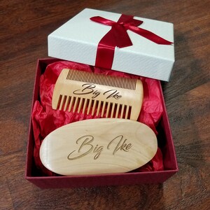 May include: A gift box with a white lid and a red ribbon contains a wooden comb and a wooden oval brush. Both items are light brown and have the engraved text "Big Ike". The box is lined with red tissue paper.
