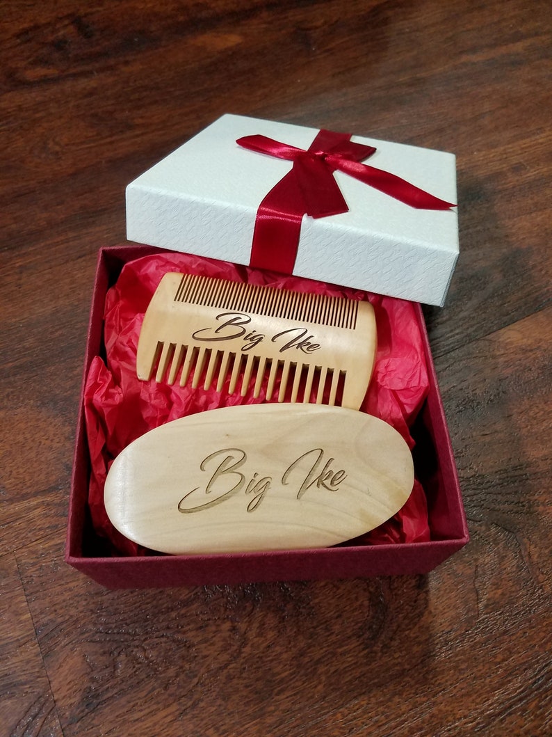 May include: A gift box with a white lid and a red ribbon contains a wooden comb and a wooden oval brush. Both items are light brown and have the engraved text "Big Ike". The box is lined with red tissue paper.