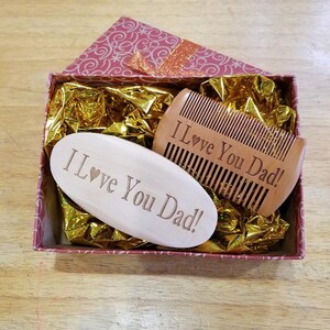 May include: A gift box with a pink and white floral pattern contains a wooden comb and a wooden oval-shaped item. Both items are engraved with the words "I Love You Dad!" The box is lined with gold-colored tissue paper.
