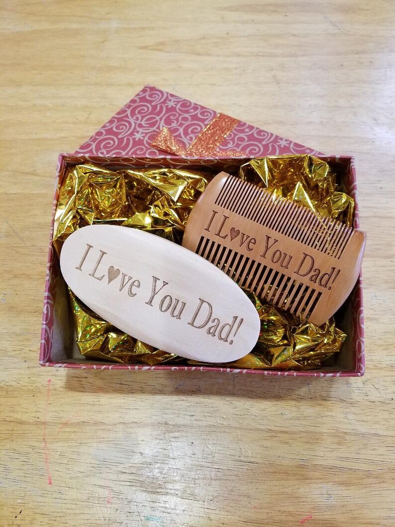 May include: A gift box with a pink and white floral pattern contains a wooden comb and a wooden oval-shaped item. Both items are engraved with the words "I Love You Dad!" The box is lined with gold-colored tissue paper.