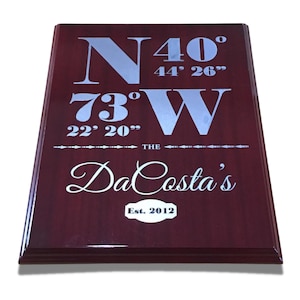 以下が含まれることがあります： 銀色の文字が書かれた濃い赤色の木製のプレート。プレートには、N40° 44' 26" 73°W 22' 20" の座標と「THE DaCosta's Est. 2012」の文字が表示されています。