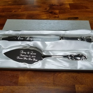 May include: A silver cake knife and server set in a white satin lined box. The knife is engraved with "Our Beginning" and the server is engraved with "Amy & Luis September 21, 2019 Forever Plus Two Days".