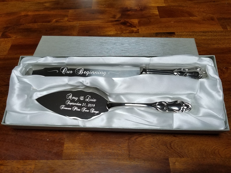 May include: A silver cake knife and server set in a white satin lined box. The knife is engraved with "Our Beginning" and the server is engraved with "Amy & Luis September 21, 2019 Forever Plus Two Days".