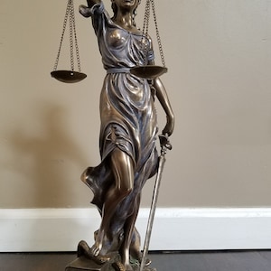 May include: A bronze statue of Lady Justice, blindfolded, holding a sword and scales. The statue is standing on a book with a snake wrapped around it. The base of the statue has a black plaque with the text "ANDREA SMITH, ESQ."