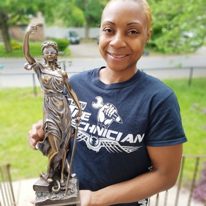 May include: A bronze statue of Lady Justice holding a scale and a sword, standing on a base with books and a snake. The statue is being held by a person wearing a black t-shirt with the text "The Technician" printed on it. The text "Dina Gattuso, Esquire" is on the base of the statue.
