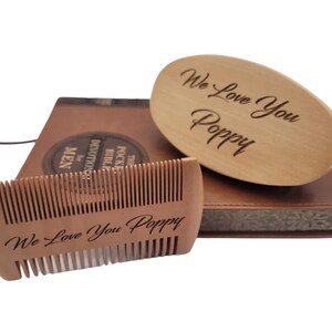 May include: A wooden beard comb and brush set with the engraved message "We Love You Poppy." The set rests on a brown leather-bound book. The comb is rectangular, and the brush is oval-shaped.